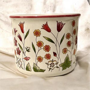 May include: A cylindrical ceramic planter with a cream-colored base and a thin red rim. The exterior features a repeating pattern of colorful wildflowers, including orange and red tulips, small yellow blossoms, and green leaves painted on the surface.