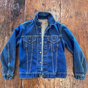 May include: A classic blue denim jacket with button closures, two chest pockets, and a collar. The jacket has a faded appearance and a red tag on the chest pocket. The jacket is displayed on a wooden surface.