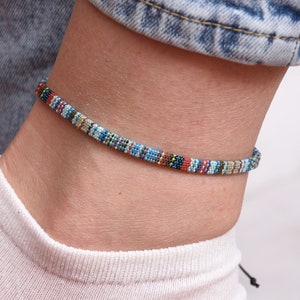 Könnte beinhalten: Ein farbenfrohes gewebtes Fußkettchen mit einem blauen, roten, gelben und weißen Muster. Das Fußkettchen ist an einem Fußgelenk einer Person zu sehen.