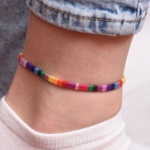 May include: A colorful woven ankle bracelet with a rainbow pattern. The bracelet is made of thin, colorful threads that are woven together to create a unique and stylish design.