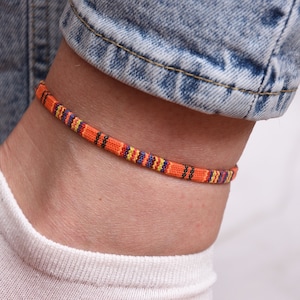 May include: A woven ankle bracelet with orange, yellow, blue and black stripes. The bracelet is worn on a person's ankle.
