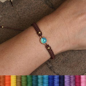 May include: A brown macrame bracelet with a central iridescent blue-green stone and gold-colored accents. The bracelet is adjustable and features a sliding knot closure. The image shows the bracelet on a wrist.