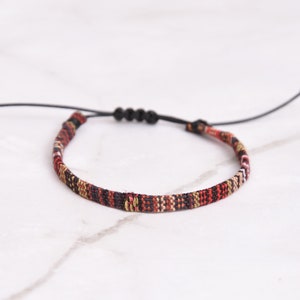 Peut inclure: Un bracelet tiss&eacute; avec un cordon noir et une fermeture r&eacute;glable. Le bracelet pr&eacute;sente un motif g&eacute;om&eacute;trique color&eacute; dans des tons de rouge, noir, blanc et or.