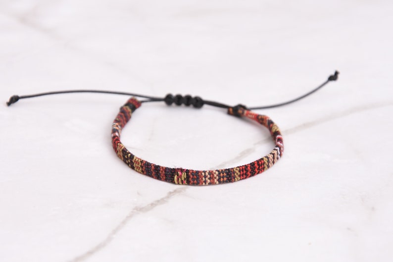 Peut inclure: Un bracelet tiss&eacute; avec un cordon noir et une fermeture r&eacute;glable. Le bracelet pr&eacute;sente un motif g&eacute;om&eacute;trique color&eacute; dans des tons de rouge, noir, blanc et or.