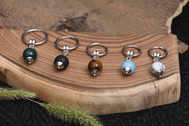 May include: Five earrings with silver hoops and gemstone beads in various colors: dark green, black, brown, light blue, and white with gray marbling. The earrings are displayed on a wooden surface.