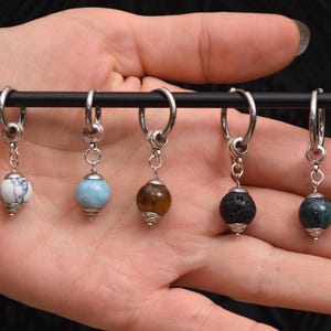 May include: A collection of six hoop earrings with different gemstone beads. The beads are white, blue, brown, black, and green. The earrings are displayed on a black bar.