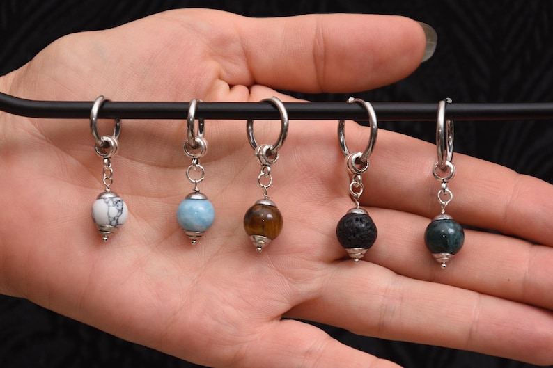 May include: A collection of six hoop earrings with different gemstone beads. The beads are white, blue, brown, black, and green. The earrings are displayed on a black bar.