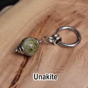 May include: A single earring with a green and white unakite stone bead and a silver-colored hoop. The earring is displayed on a light brown wooden surface. The word "Unakite" is written in white text.