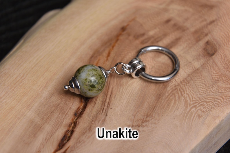 May include: A single earring with a green and white unakite stone bead and a silver-colored hoop. The earring is displayed on a light brown wooden surface. The word "Unakite" is written in white text.