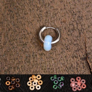 Puede incluir: Un pendiente de aro de plata con una perla de ópalo azul claro. El pendiente se muestra sobre un fondo de madera marrón. La imagen también muestra una variedad de otras cuentas en diferentes colores y materiales.