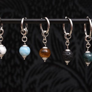 May include: Five silver hoop earrings with dangling gemstone beads. The beads are white, light blue, amber, black, and green. The earrings are displayed against a dark, textured background.