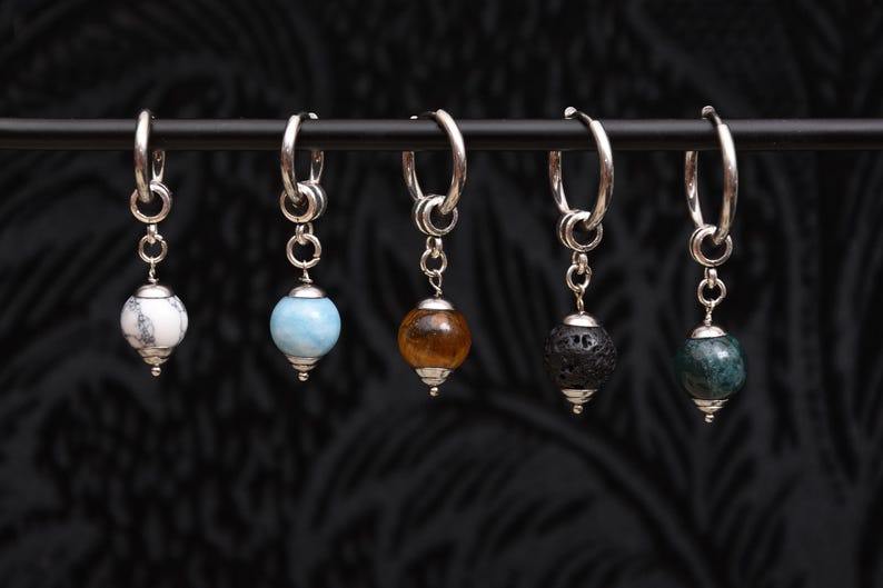 May include: Five silver hoop earrings with dangling gemstone beads. The beads are white, light blue, amber, black, and green. The earrings are displayed against a dark, textured background.