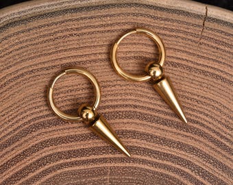 May include: A pair of gold-colored hoop earrings with a conical spike pendant. The earrings are displayed on a brown, textured surface. The earrings have a simple, yet bold design.