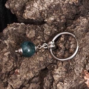 May include: A keychain with a dark teal, spherical bead and a silver-colored ring. The bead is attached to the ring with a small chain. The background is a piece of rough, brown bark.