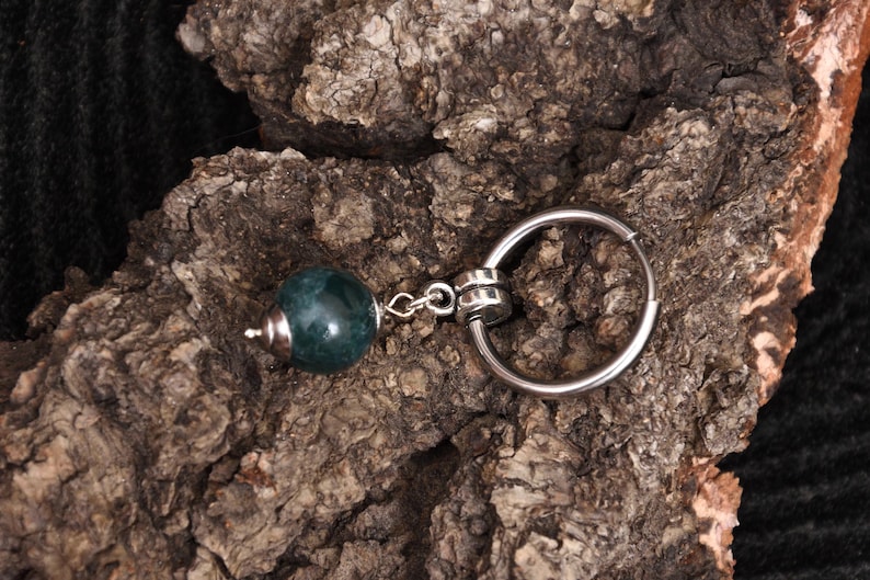 May include: A keychain with a dark teal, spherical bead and a silver-colored ring. The bead is attached to the ring with a small chain. The background is a piece of rough, brown bark.