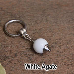 May include: A white agate charm with a silver-colored metal ring and a small, dangling white bead. The charm is displayed on a textured, brown surface. The text "White Agate" is at the bottom.