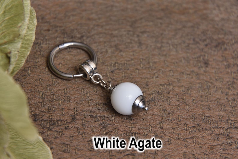 May include: A white agate charm with a silver-colored metal ring and a small, dangling white bead. The charm is displayed on a textured, brown surface. The text "White Agate" is at the bottom.
