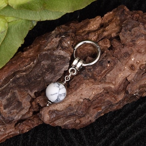 May include: A silver-toned earring with a white and gray marbled stone bead. The earring is displayed on a piece of textured brown bark, with green leaves in the background. The earring has a hoop design.