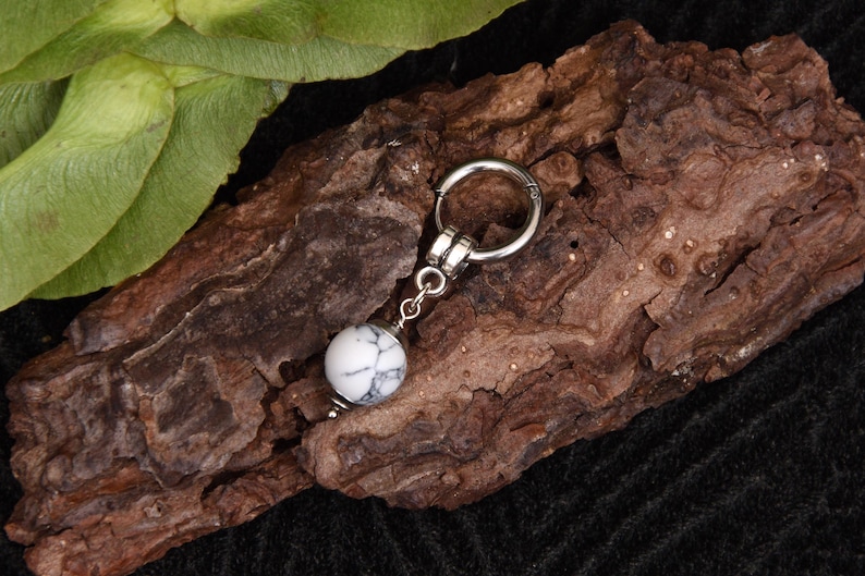 May include: A silver-toned earring with a white and gray marbled stone bead. The earring is displayed on a piece of textured brown bark, with green leaves in the background. The earring has a hoop design.