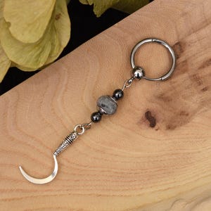 Pode incluir: Um amuleto de lua crescente prateado com uma corrente de contas presa a um brinco de argola prateado. As contas são pretas e cinzas.