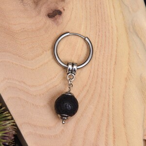 May include: A single silver hoop earring with a black lava stone bead pendant. The earring is displayed on a light-colored wooden surface. The lava stone bead is round and textured.