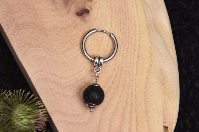 May include: A single silver hoop earring with a black lava stone bead pendant. The earring is displayed on a light-colored wooden surface. The lava stone bead is round and textured.