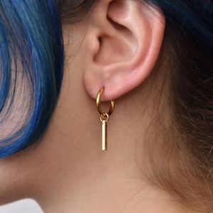 Puede incluir: Pendiente de aro dorado con un colgante de barra vertical. El pendiente se muestra en una oreja, con cabello azul en el fondo. Diseño simple y minimalista.