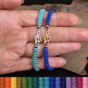 May include: Two macrame bracelets, one turquoise and one blue, each featuring a treble clef charm. The turquoise bracelet has a silver charm, while the blue bracelet has a gold charm. Both bracelets are displayed on a hand.