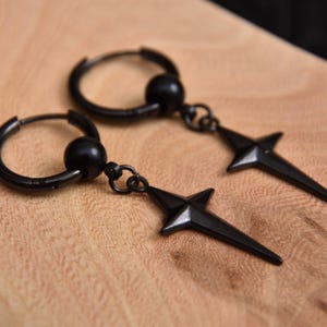 Op de afbeelding: Een paar zwarte oorringen met stervormige hangers. De oorbellen zijn van metaal en liggen op een houten oppervlak. De sterhangers hebben puntige randen en zijn aan de ringen bevestigd.