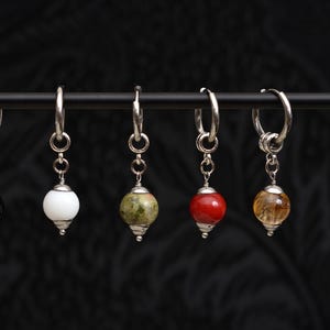 May include: Six silver hoop earrings with gemstone pendants. The pendants are various colors, including black, white, green, red, yellow, and blue. The earrings are displayed against a dark, textured background.