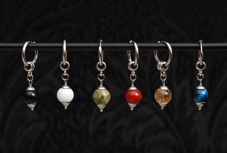 May include: Six silver hoop earrings with gemstone pendants. The pendants are various colors, including black, white, green, red, yellow, and blue. The earrings are displayed against a dark, textured background.