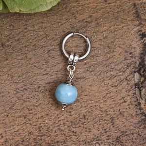 May include: A single silver hoop earring with a light blue, round bead pendant. The earring is set against a textured, brown background. The bead is approximately 0.5 inches in diameter.
