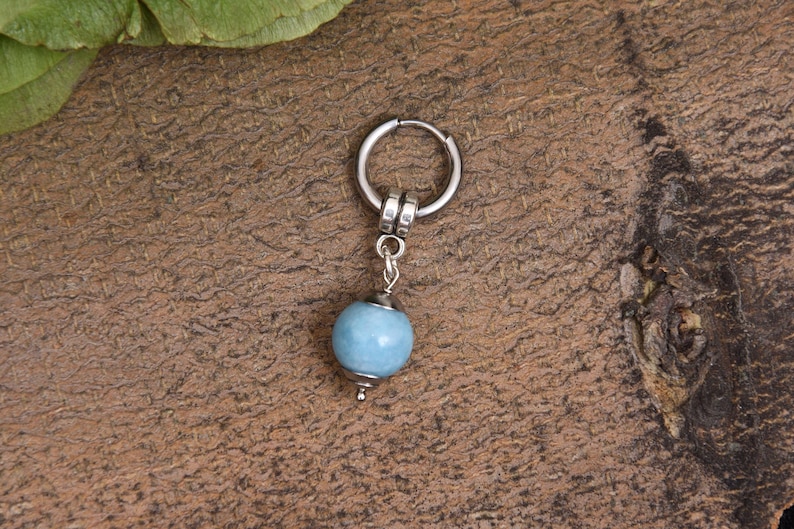 May include: A single silver hoop earring with a light blue, round bead pendant. The earring is set against a textured, brown background. The bead is approximately 0.5 inches in diameter.