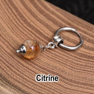 May include: A citrine gemstone charm with a silver-colored metal ring. The gemstone is a light orange color and has a silver-colored metal cap. The word "Citrine" is written in white text.