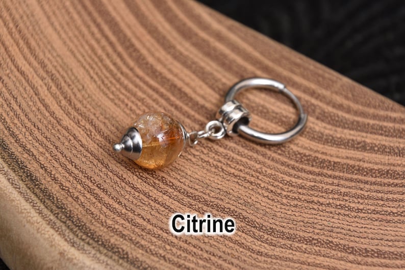 May include: A citrine gemstone charm with a silver-colored metal ring. The gemstone is a light orange color and has a silver-colored metal cap. The word "Citrine" is written in white text.