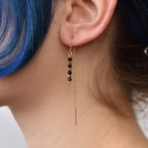 Könnte beinhalten: Goldfarbene Ohrhänger mit schwarzen Perlen. Die Ohrringe haben eine zarte Kette mit kleinen, runden schwarzen Perlen in der Nähe des Ohrs. Das Design ist schlicht und elegant. Ein stilvolles Accessoire.