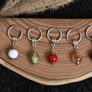 Puede incluir: Seis llaveros con aros plateados y colgantes de piedras preciosas en negro, blanco, verde, rojo, ámbar y azul. Los llaveros se exhiben sobre una superficie de madera.