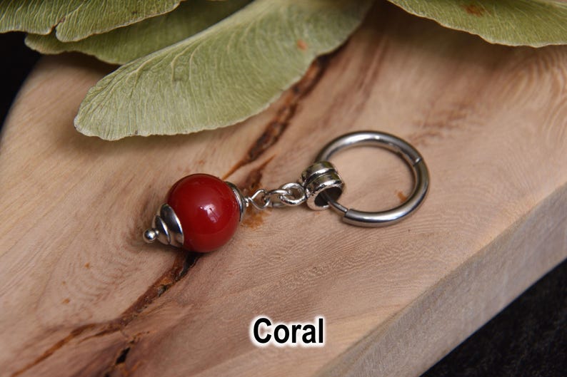 May include: A single earring with a red coral bead and a silver-colored hoop. The coral bead has a shiny finish and a silver-colored conical cap. The word "Coral" is written in white text at the bottom of the image.