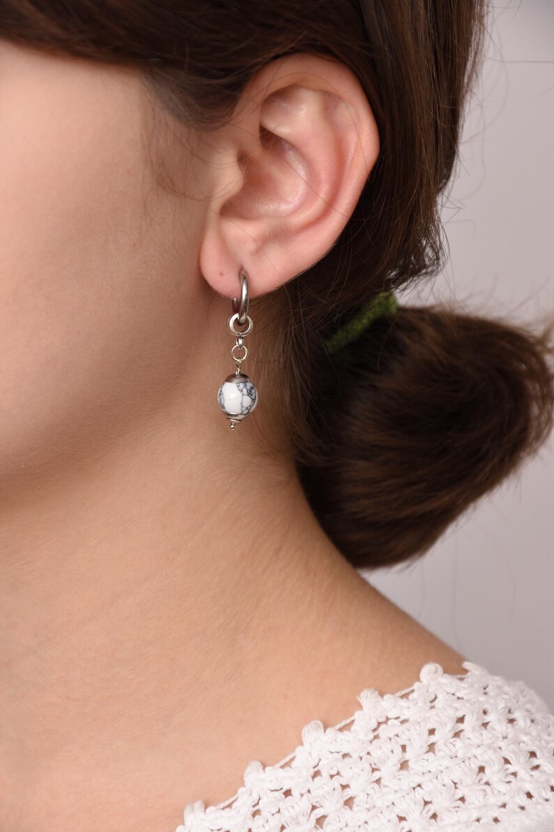 May include: A silver hoop earring with a dangling white and black marbled bead. The earring is attached to a small silver chain. The earring is shown on a person's ear.