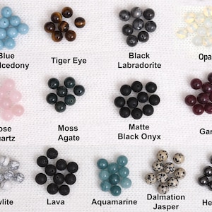 May include: A collection of gemstone beads in various colors and types, including blue chalcedony, tiger eye, black labradorite, and opal. Other stones include rose quartz, moss agate, matte black onyx, and garnet. Additional beads are howlite, lava, aquamarine, dalmatian jasper, and hematite.