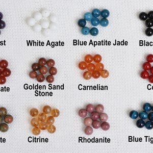 May include: A collection of gemstone beads in various colors, including amethyst, white agate, blue apatite jade, and black agate. Other stones include red agate, golden sand stone, carnelian, coral, unakite, citrine, rhodonite, and blue tiger eye. Each stone is labeled.