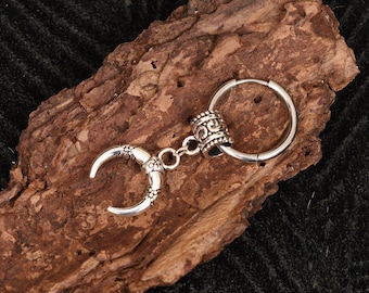 May include: A silver-colored earring with a crescent-shaped charm and a decorative cylindrical bead. The earring is attached to a circular hoop. The charm has a detailed, textured design. The earring is displayed on a piece of brown wood.