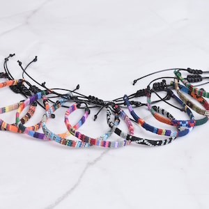 Peut inclure: Un ensemble de 12 bracelets tiss&eacute;s color&eacute;s avec des cordons noirs r&eacute;glables. Les bracelets pr&eacute;sentent divers motifs g&eacute;om&eacute;triques et des couleurs vives, notamment le rouge, l'orange, le jaune, le vert, le bleu, le violet et le blanc.