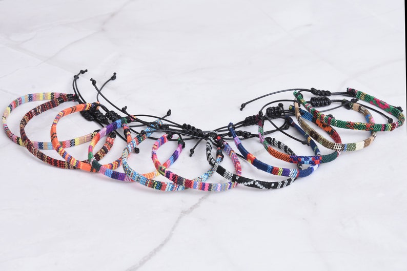 Peut inclure: Un ensemble de 12 bracelets tiss&eacute;s color&eacute;s avec des cordons noirs r&eacute;glables. Les bracelets pr&eacute;sentent divers motifs g&eacute;om&eacute;triques et des couleurs vives, notamment le rouge, l'orange, le jaune, le vert, le bleu, le violet et le blanc.