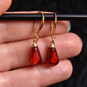May include: A pair of gold earrings featuring a teardrop-shaped, deep red gemstone pendant. The earrings hang from gold hooks. The background is a dark, blurred texture.