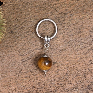 May include: A silver-colored keychain with a round, polished tiger's eye gemstone pendant. The gemstone is a warm brown color with darker brown and black streaks. The keychain is suspended from a silver ring.