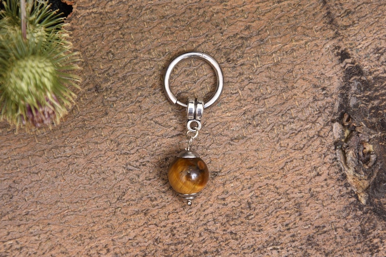 May include: A silver-colored keychain with a round, polished tiger's eye gemstone pendant. The gemstone is a warm brown color with darker brown and black streaks. The keychain is suspended from a silver ring.