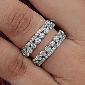 Puede incluir: Dos anillos de plata con piedras preciosas transparentes. Uno presenta una fila de gemas redondas, el otro una doble fila de piedras más pequeñas. Los anillos están en una mano, con una manga burdeos en el fondo.