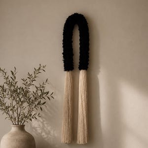 以下が含まれることがあります： 黒いテクスチャーのあるアーチ型の壁掛けで、両端から長いベージュのタッセルが垂れ下がっています。この装飾品は、『Kinfolk』、『AD at 100』、『Chanel』というタイトルの3冊の本の積み重ねの上に飾られており、横にはベージュの花瓶と火のついたキャンドルが置かれています。