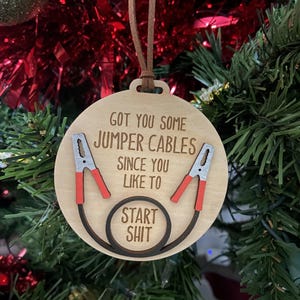 Puede incluir: Un adorno navideño de madera con forma de círculo y un cordón de cuero marrón. El adorno tiene el texto "Got you some jumper cables since you like to start shit" con una ilustración roja y negra de cables de arranque.